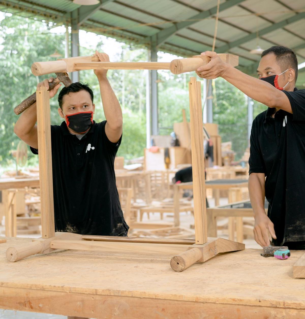 Inside an ordinary UK dining room, two self-employed delivery crew members assembling a wooden dining table, one aligning…